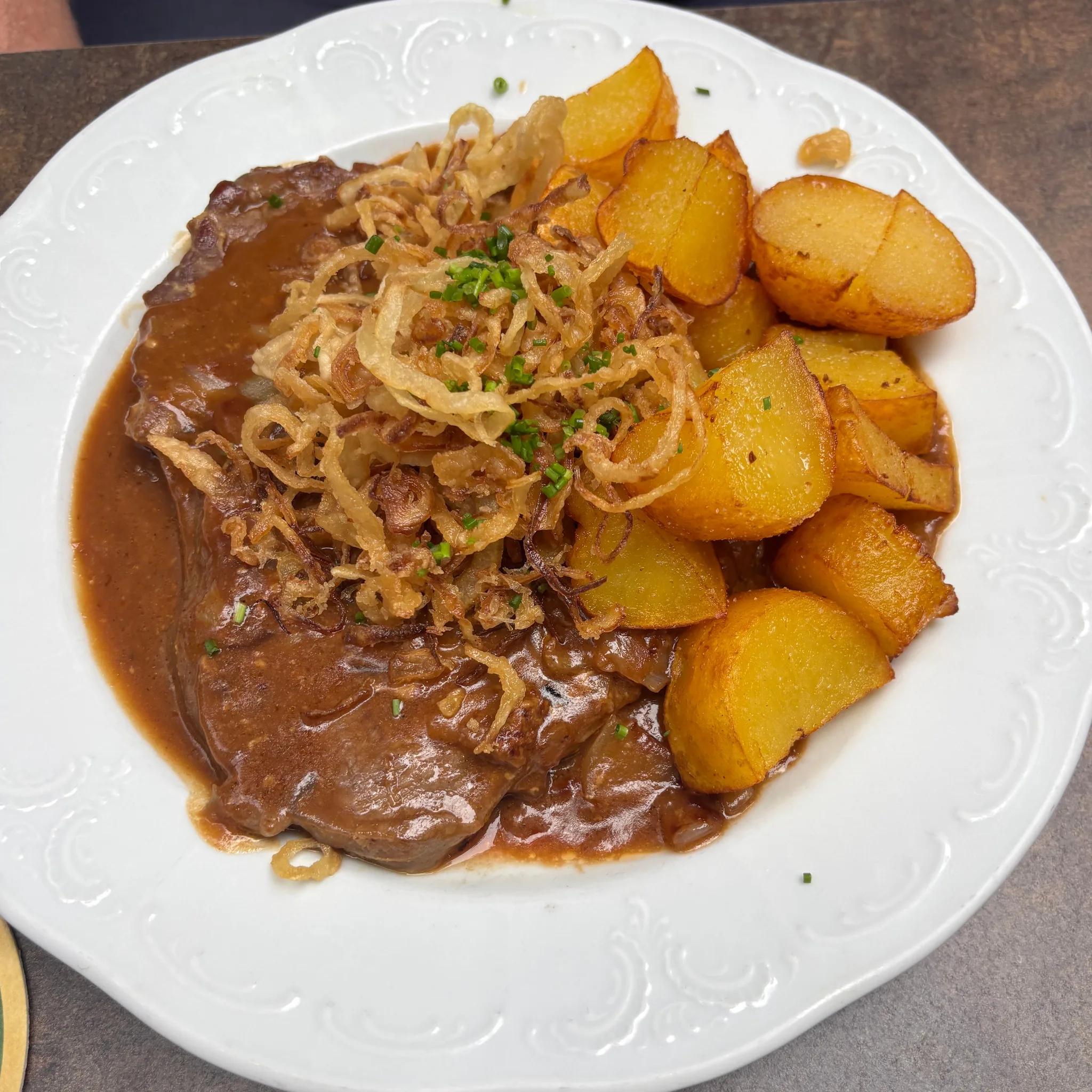 Rostbraten mit Röstzwiebeln und Braterdäpfeln auf einem weißen Teller mit Reliefrand, fotografiert von einem Gast