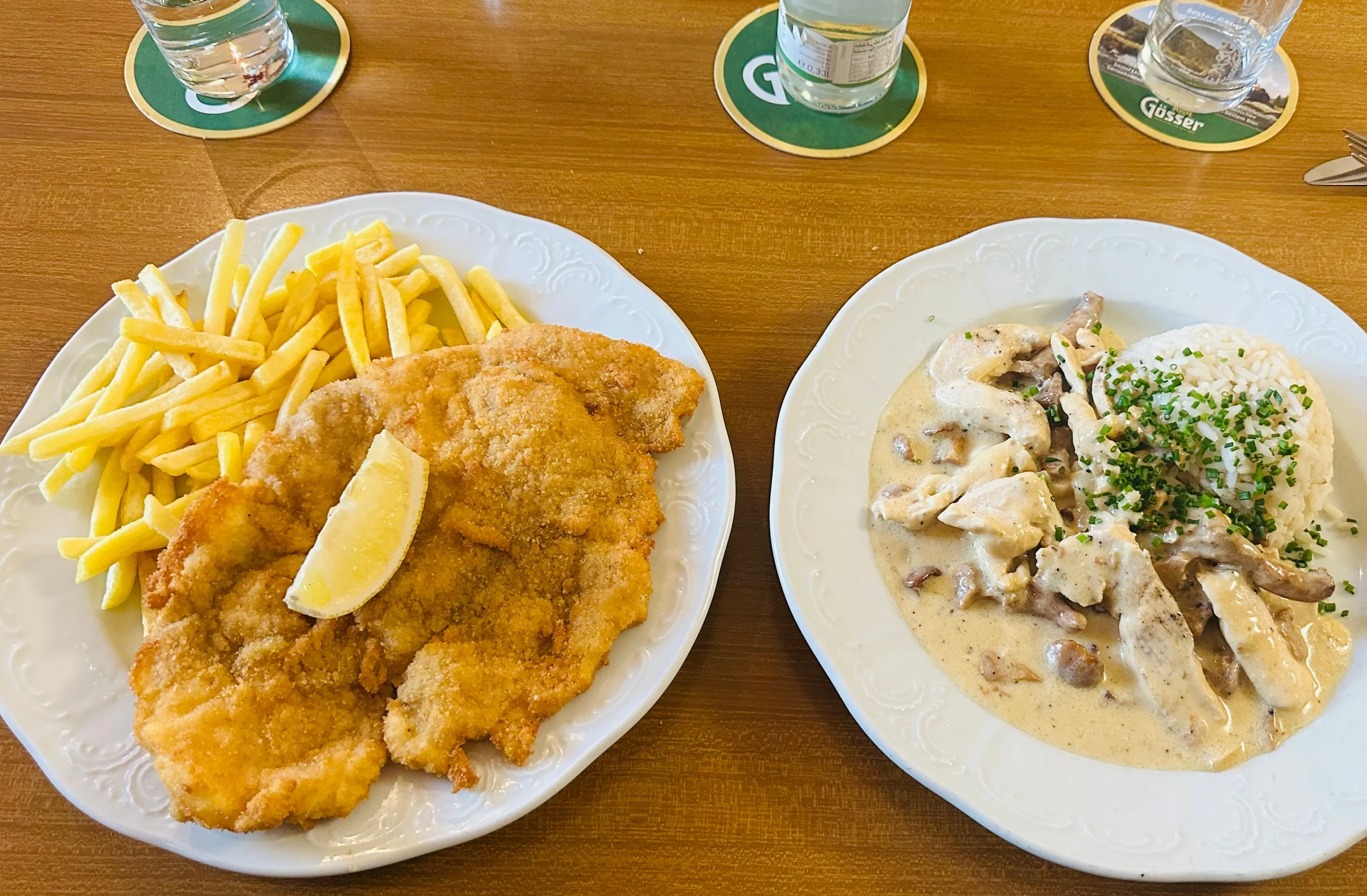 Wirtshaustisch von oben: ein Schnitzel mit Pommes frites und Zitrone, daneben ein Rahmgericht mit Reis, Gläser auf Bierdeckeln